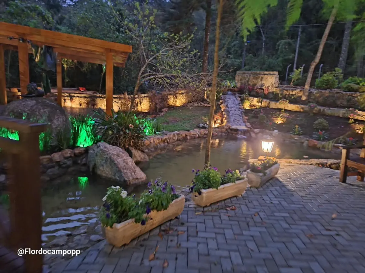Jardim iluminado à noite com lago, muro de pedra, pergolado de madeira e piso intertravado